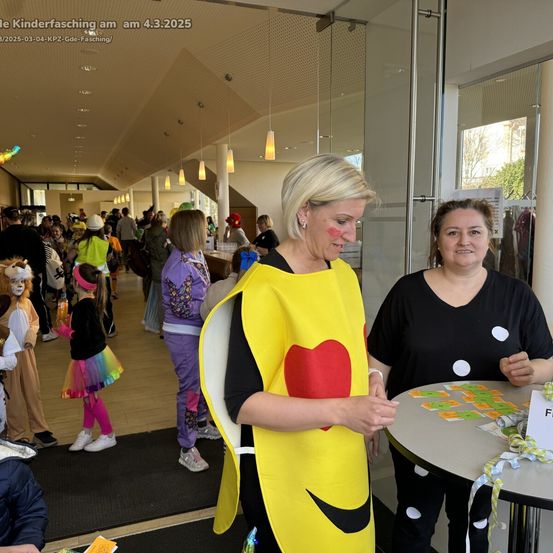 Eine Frau in einer Bananen-Kostüm steht an einem Tisch mit einer anderen Frau. Dahinter steht eine Menge Leute, einige in Kostümen. Das Datum ist der 4. März 2025.