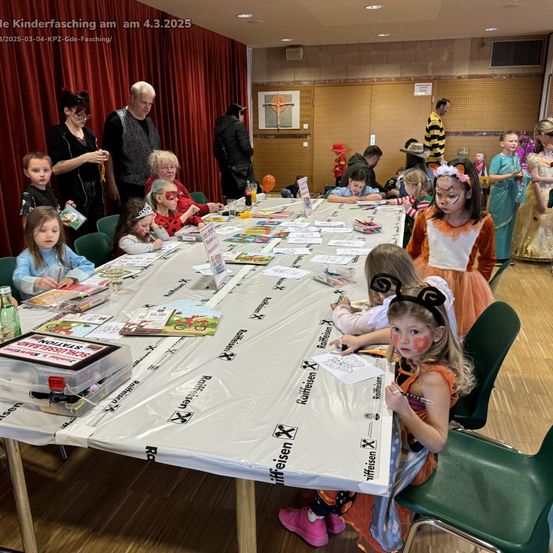 Kinder in Kostümen sitzen um einen Tisch mit Büchern und Markern herum. Ein Erwachsener steht hinter dem Tisch. Sie befinden sich wahrscheinlich auf einem Kinderkarneval.