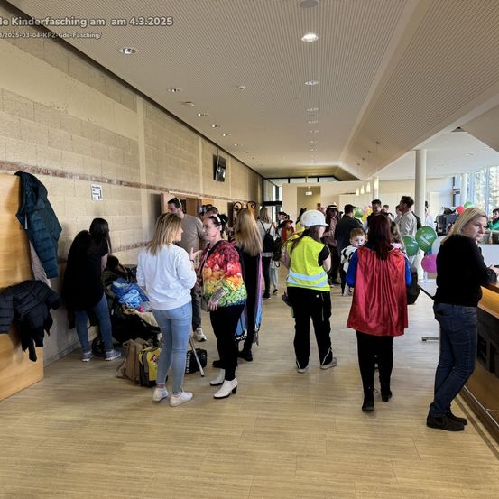 Eine Gruppe von Menschen befindet sich in einem Flur eines Gebäudes mit Luftballons und Mänteln, die an der Wand hängen. Eine Frau in einer gelben Weste schaut jemanden an. Ein Kinderwagen mit Taschen ist in der Nähe.