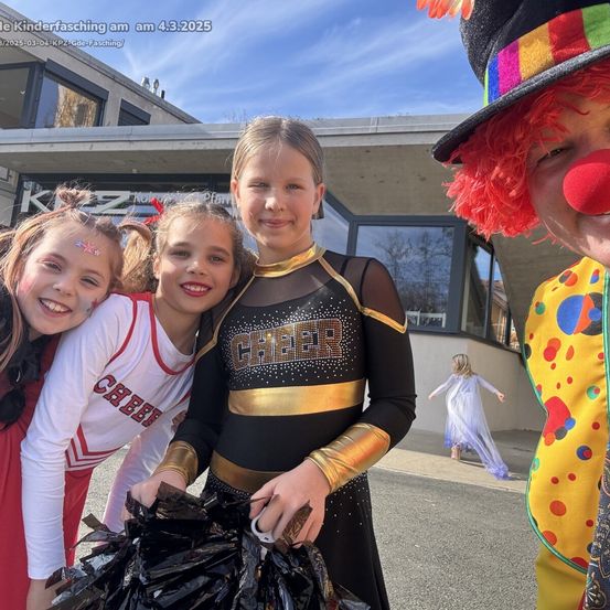 Drei Mädchen in Cheerleader-Uniformen, eines mit einem Pom-Pom, und ein Clown neben ihnen vor einem Gebäude.