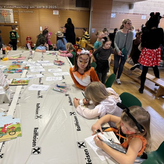 Ein Raum mit Kindern in Kostümen. Sie sitzen an einem langen Tisch mit Malbüchern und Buntstiften. Im Hintergrund stehen und sitzen weitere Kinder. Es gibt ein Schild, das Kinderfasching besagt.