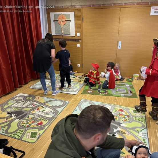 Kinder in Kostümen spielen auf Matten in einem Raum mit einem Kruzifix. Erwachsene beobachten und interagieren. Datum und Veranstaltungsname sind auf dem Bild.