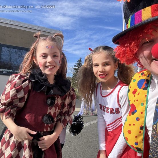 Zwei junge Mädchen in Kostümen posieren mit einem Clown, alle lächeln. Im Hintergrund ist ein Gebäude und ein klarer Himmel zu sehen. Der Clown trägt einen bunten Hut, und ein Mädchen hat Gesichtsbemalung.