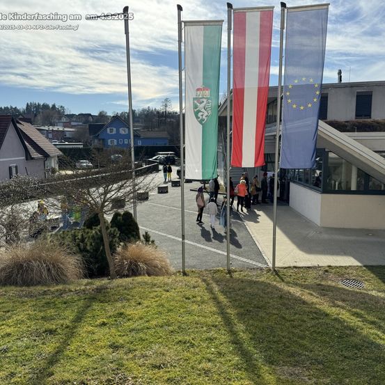 Außenbereich mit Fahnen vor einem Gebäude, Menschen gehen und ein blauer Himmel mit Wolken.