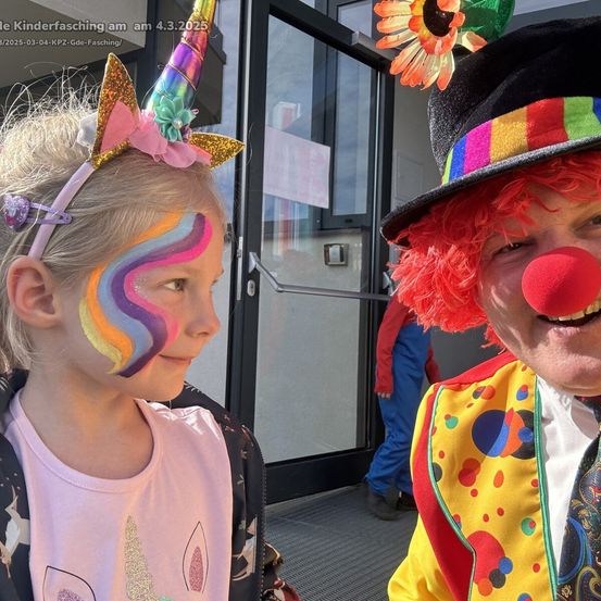 Ein junges Mädchen mit einem Einhorn-Kopfband und Regenbogen-Gesichtsbemalung steht neben einem Clown mit roter Nase.
