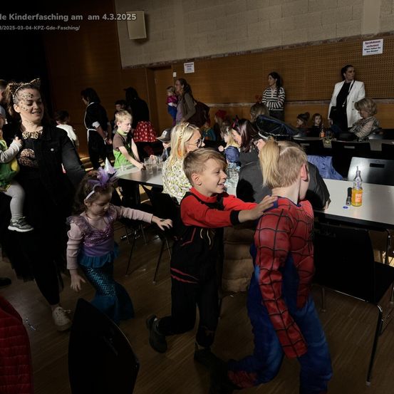 Eine Gruppe von Kindern und Erwachsenen versammelt sich in einer Halle für eine Kostümparty. Die Kinder sind in verschiedenen Kostümen gekleidet, darunter eine Meerjungfrau, eine Katze und ein Superheld. Die Erwachsenen beobachten, wie die Kinder spielen und miteinander interagieren.