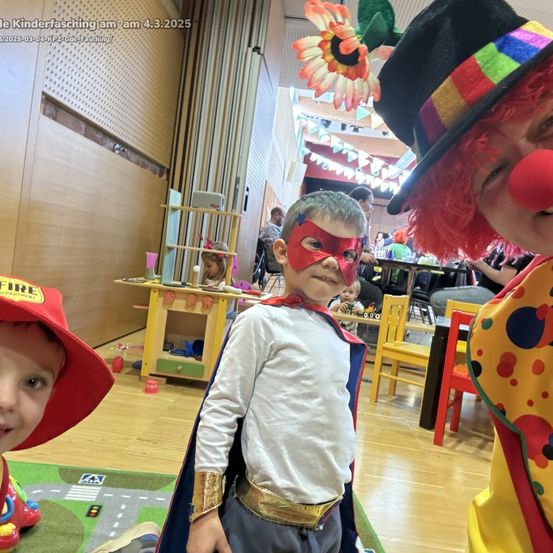 Drei Kinder sind verkleidet, eines mit einem Feuerwehrmannhut, ein anderes in einem Superheldenkostüm und ein Clown in der Nähe. Die Umgebung ist drinnen mit bunten Dekorationen und Holzboden.