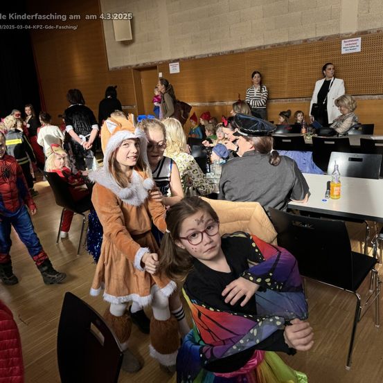 Eine Gruppe von Kindern in verschiedenen Kostümen versammelt sich in einem Saal, möglicherweise für eine Feier. Einige tragen Brillen, Hüte und Schuhe, während andere auf Stühlen sitzen. Eine Frau steht hinter einem Tisch mit Flaschen.