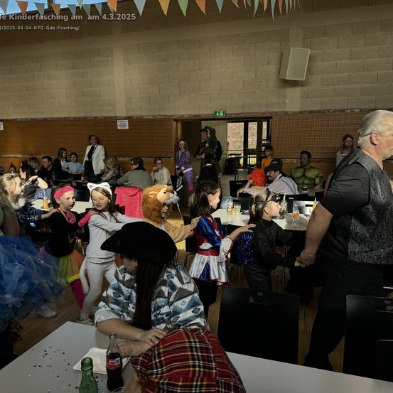 Eine große Gruppe von Menschen in Kostümen versammelt sich in einem Saal für eine Feier. Mehrere Kinder tanzen, während andere an Tischen sitzen. Auf den Tischen stehen verschiedene Getränke, und ein Löwenkostüm ist in der Mitte zu sehen.
