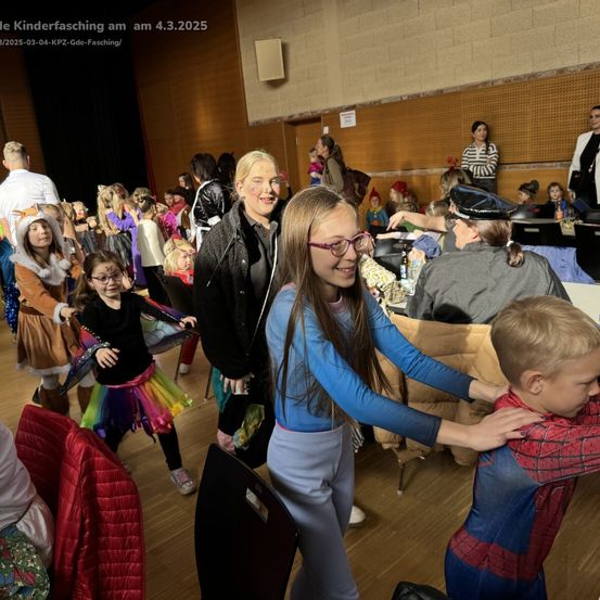 Eine Gruppe von Kindern und Erwachsenen befindet sich in einem Saal, möglicherweise für einen Kinderfasching. Sie tragen verschiedene Kostüme. Ein Mädchen mit Brille hält einen Jungen in einem Spider-Man-Kostüm.