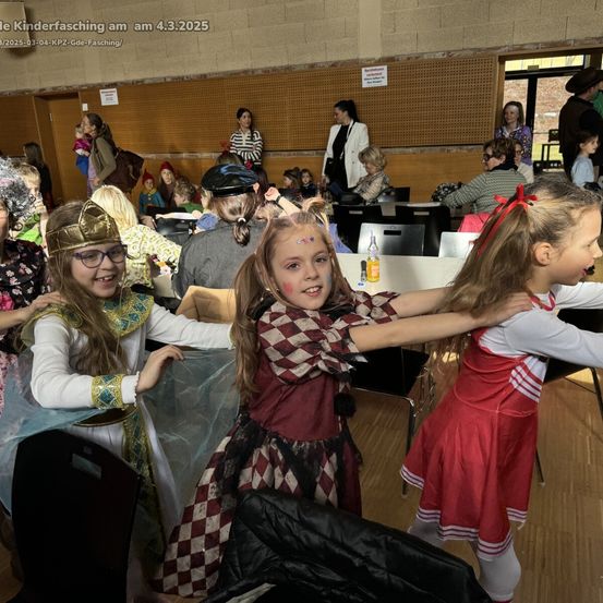 Ein Kinderfasching am 4. März 2025 zeigt Kinder in Kostümen. Ein Mädchen in einem roten Kleid tanzt mit einer anderen, während Erwachsene von Stühlen aus zusehen. Tische und Stühle sind aufgestellt.