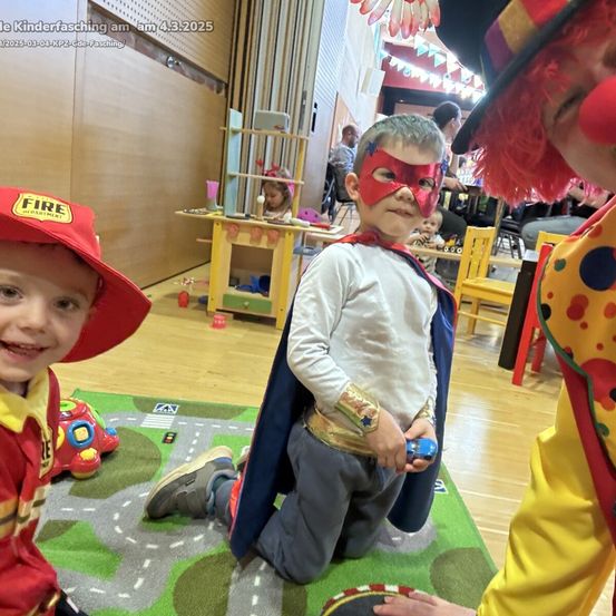 Drei Kinder in Kostümen, eines als Feuerwehrmann, ein anderes als Superheld und ein Clown. Sie spielen auf einem Teppich in einem Raum mit Spielzeug und Stühlen.