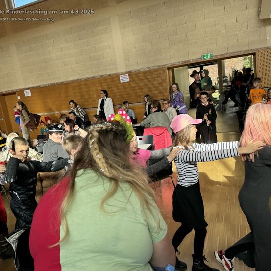 Menschen versammeln sich in Innenräumen für einen Kinderfasching am 4.3.2025. Mehrere Kinder sind in Kostümen gekleidet, und zwei Mädchen umarmen sich in der Mitte.