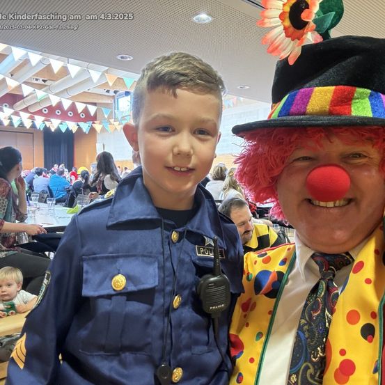 Ein junger Junge in einer Polizeiuniform steht neben einem Clown mit einer roten Nase bei einer Veranstaltung.