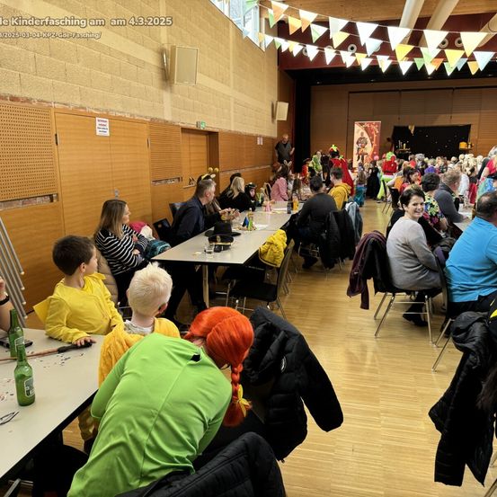 Eine Gruppe von Menschen sitzt an Tischen in einer Halle. Sie tragen Kostüme und einige halten Essen. Der Saal hat Holzböden und Wände mit Dekorationen. Es stehen auch Menschen im Hintergrund.