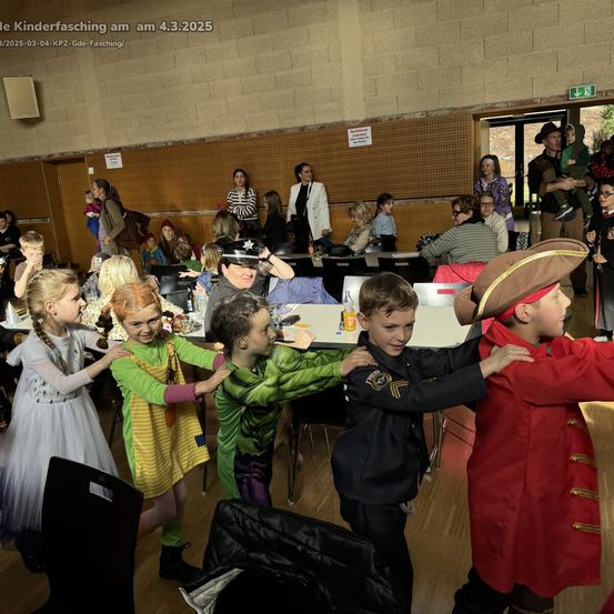 Eine Gruppe von Kindern in Kostümen tanzt in einem Raum mit Tischen und Stühlen. Einige tragen Brillen und Hüte. Erwachsene beobachten.