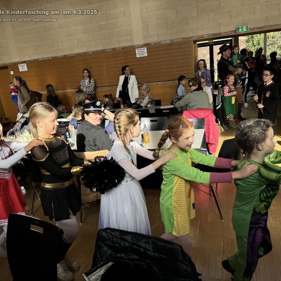 Eine Gruppe von Kindern in verschiedenen Kostümen tanzt in einem Saal, während Erwachsene zusehen. Ein Banner im Hintergrund lautet 'Kinderfasching am 4.3.2025'.