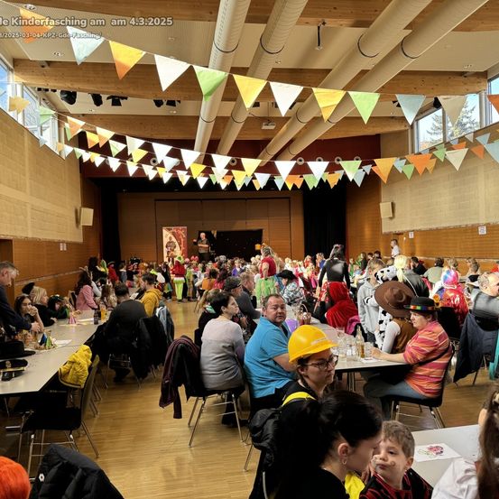 Eine große Indoor-Veranstaltung, möglicherweise ein Kinderkarneval, mit bunten Wimpelketten. Menschen sitzen an Tischen, einige in Kostümen, und zwei Personen stehen in der Nähe der Bühne. Der Raum hat Holzböden und große Fenster.