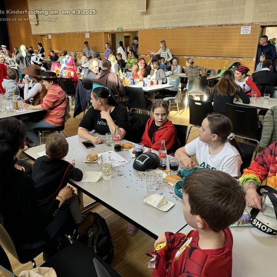 Ein Saal voller Menschen in Kostümen, die um Tische herum sitzen. Einige Kinder tragen Superheldenkostüme. Die Tische sind mit Essen, Getränken und Papieren gedeckt.