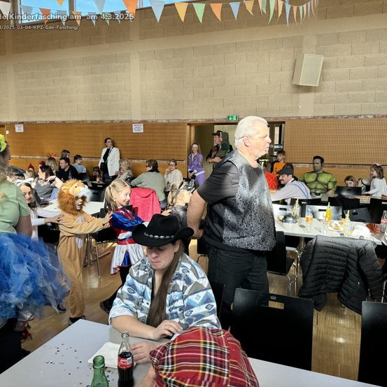 Eine große Gruppe von Menschen ist drinnen versammelt, einige in Kostümen, bei einer Veranstaltung. Tische sind mit verschiedenen Gegenständen besetzt, und ein Mann in einem schwarzen Cowboyhut schaut eine Frau an, die vor ihm sitzt.