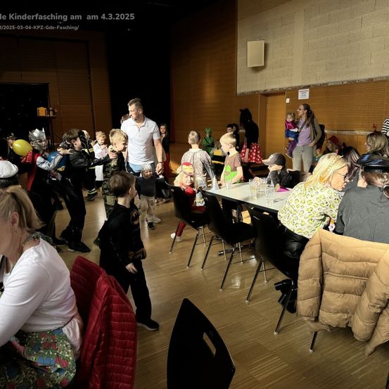 Eine Gruppe von Kindern in Kostümen versammelt sich in einem Raum mit Erwachsenen und Tischen, eines hält einen Ballon.