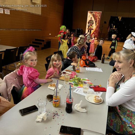 Eine Gruppe von Kindern und Erwachsenen sitzt an einem langen Tisch, in Kostümen gekleidet, wahrscheinlich für einen Kinderfasching. Sie sind von verschiedenen Essensartikeln umgeben, einschließlich eines Donuts. Dahinter ist eine Bühne aufgebaut, mit einem Hintergrund, der auf eine bevorstehende Veranstaltung hindeutet.