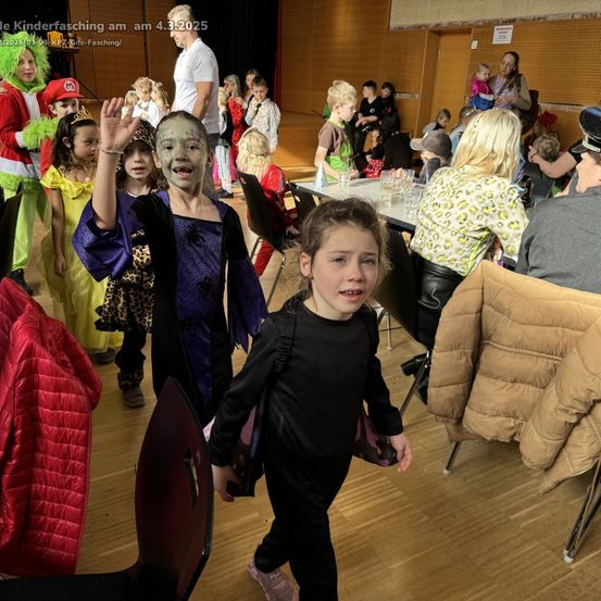 Kinder in Kostümen, darunter eine Hexe und ein Pirat, versammeln sich für eine Feier. Einige sitzen, andere stehen, und Erwachsene sind anwesend. Tische mit Gegenständen und Stühlen sind aufgestellt.