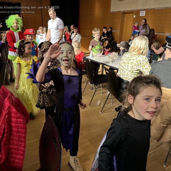 Kinder in Kostümen versammeln sich am 4. März 2025 zu einer Feier. Ein Kind winkt begeistert, während andere hinter einem Tisch mit Gläsern stehen.