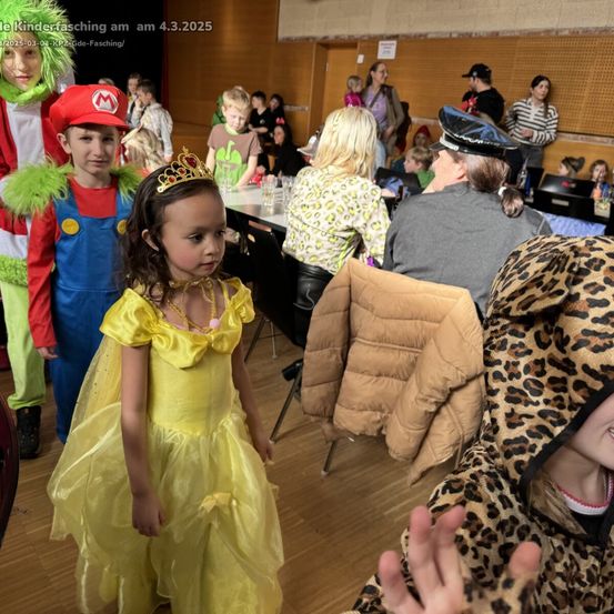 Eine Gruppe von Kindern in Kostümen, darunter Mario, eine Prinzessin und ein Leopard, steht in einem Raum mit Erwachsenen, die an Tischen sitzen. Die Umgebung deutet auf eine Kostümparty oder ein thematisches Ereignis hin.