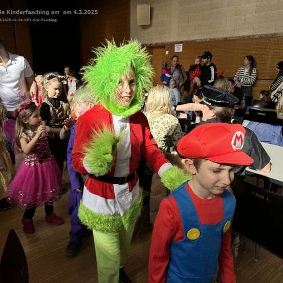 Eine Gruppe von Kindern und Erwachsenen ist in einem Saal, einige sind in Kostümen. Ein Junge in einem Grinch-Kostüm hält die Hand eines Jungen in einem Mario-Kostüm. Andere Kinder sind ebenfalls in Kostümen. Im Hintergrund befinden sich Tische mit Stühlen.