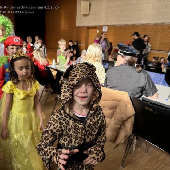 Eine Gruppe von Kindern in verschiedenen Kostümen steht in einem Saal. Ein Kind in einem Leoparden-Kostüm schaut zur Kamera. Andere Kinder in Kostümen stehen hinter ihnen. Einige Erwachsene sitzen auf Stühlen.