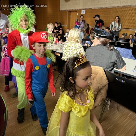 Eine Gruppe von Kindern in Kostümen, die durch einen überfüllten Raum gehen, einige Erwachsene sitzen an Tischen, ein Wasserzeichen in der oberen linken Ecke des Bildes.