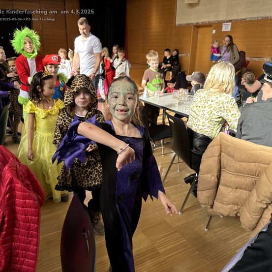 Eine Gruppe von Kindern in Kostümen, darunter eine Hexe und eine Prinzessin, steht in einem Raum mit Erwachsenen und Tischen, möglicherweise bei einer Kinderparty.