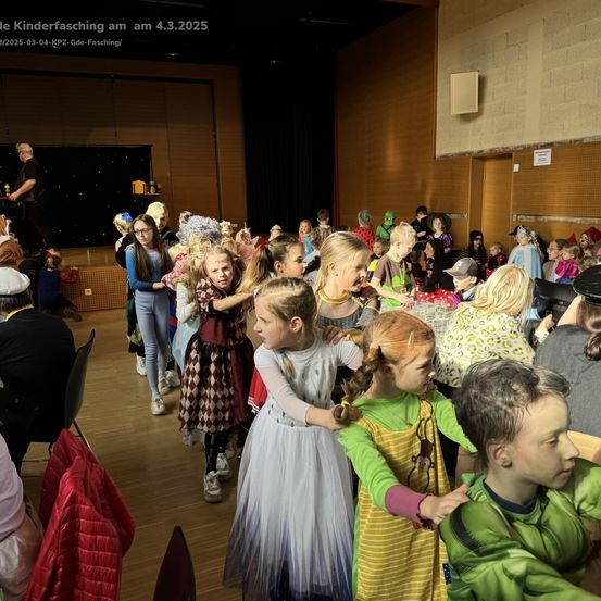 Eine Gruppe von Kindern in verschiedenen Kostümen, möglicherweise für eine Schulveranstaltung, steht in einem Saal. Der Saal hat Holzwände und eine Bühne im Hintergrund. Einige Kinder halten Händchen, während andere allein stehen.