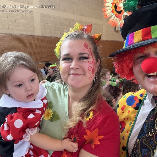 Eine Frau mit Gesichtsbemalung hält ein Kind in einem roten Pünktchenkleid, neben einem Clown mit roter Nase und buntem Hut. Die Umgebung scheint ein Karneval oder ähnliches Ereignis zu sein.