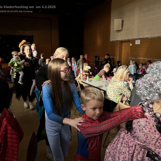 Eine Gruppe von Kindern und Erwachsenen versammelt sich in einem Saal für eine Kostümparty. Einige sind in Kostümen gekleidet und andere sitzen auf Stühlen. Es gibt einen Tisch mit Gläsern und anderen Gegenständen darauf.