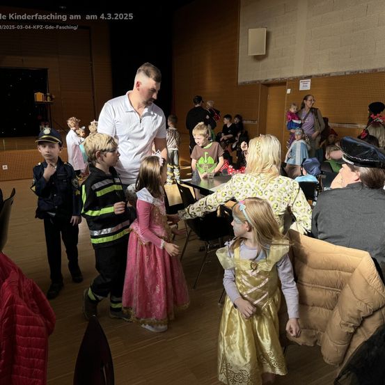 Eine Gruppe von Kindern in Kostümen und Erwachsenen bei einer Kostümparty, wobei ein Mann die Kinder auf der Bühne führt.