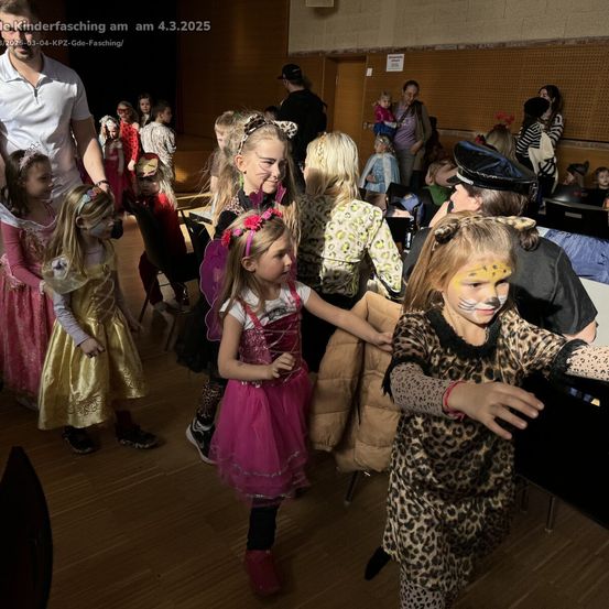 Mehrere Kinder in Kostümen tanzen in einem Saal. Einige sind als Katzen, andere als Prinzessinnen gekleidet. Eine Frau in einem Hut schaut sie an. Ein Mann in einem weißen Hemd steht in der Nähe der Kinder.