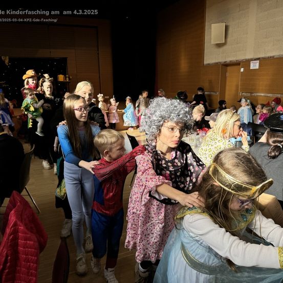 Eine Gruppe von Kindern und Erwachsenen in Kostümen versammelt sich in einem Raum, möglicherweise für eine Kostümparty. Ein Kind, das als Spider-Man verkleidet ist, wird von einer Frau umarmt.
