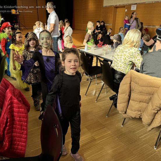 Eine Gruppe von Kindern in Kostümen bei einem Kinderfasching, einige sitzen an Tischen mit Getränken. Ein erwachsener Mann steht hinter ihnen, und eine Frau mit einem Kind auf dem Schoß sitzt an einem Tisch.