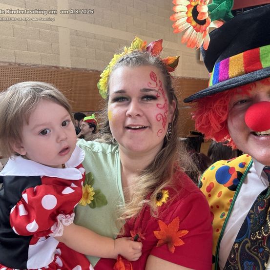 Eine Frau mit Gesichtsbemalung hält ein junges Mädchen in einer Minnie-Maus-Kostüm, mit einem Clown in einem mehrfarbigen Hut in der Nähe. Sie lächeln für ein Foto.