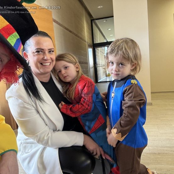 Eine Frau in einem weißen Mantel posiert mit zwei Kindern in Kostümen, eines in einem Spiderman-Outfit und das andere in einem blau-braunen Kostüm. Das Bild enthält einen Wasserzeichen mit dem Datum 4.3.2025.