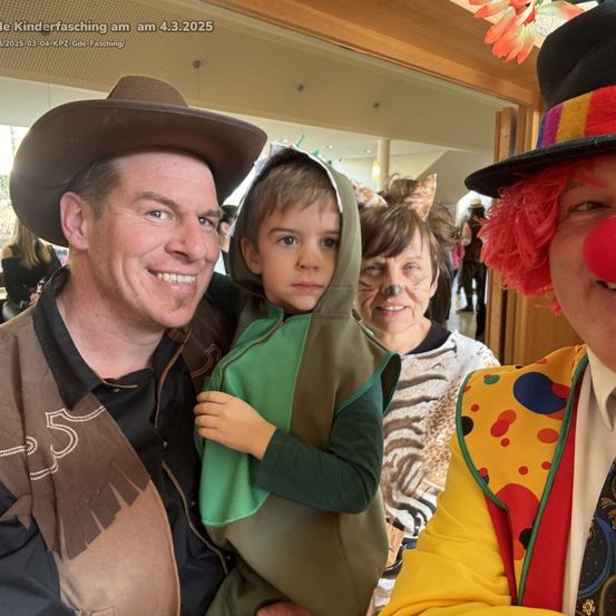 Ein Foto eines Mannes in einem Cowboyhut und eines kleinen Jungen in einem grünen Kostüm, die lächeln. Dahinter eine Frau in einem Zebrakostüm und ein Clown mit roten Haaren und einer roten Nase. Das Foto ist vom 4.3.2025.