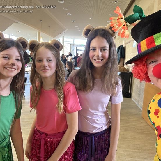 Vier Mädchen in Kostümen stehen eng beieinander in einem Saal. Die Mädchen tragen Tierkopfbedeckungen und das Mädchen rechts trägt Clowns-Make-up. Es gibt einen Wasserzeichen oben links.