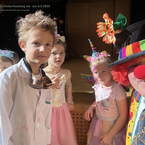 Eine Gruppe von Kindern und ein Erwachsener in Clown-Kostüm stehen zusammen in einer farbenfrohen Umgebung. Die Kinder sind in rosa Kleidern und verspielten Hüten gekleidet. Der Clown hat eine rote Nase und einen mehrfarbigen Hut.