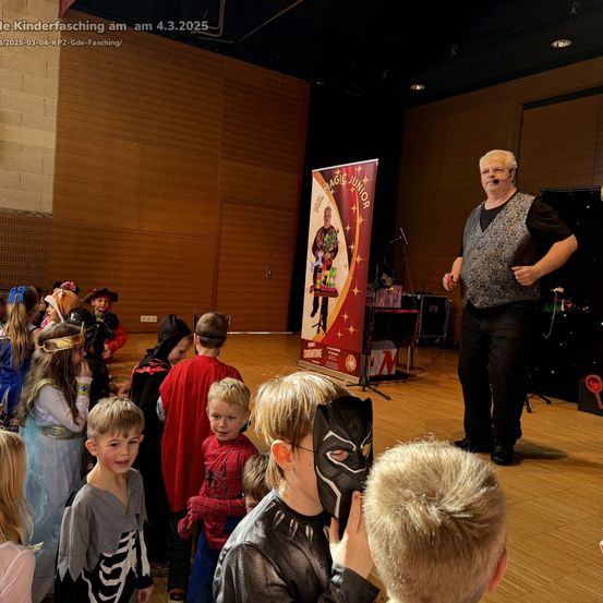 Eine Gruppe von Kindern in Kostümen versammelt sich um einen Magier auf der Bühne, mit einem Hintergrund, der 'Magic Junior' steht. Der Magier hält ein Mikrofon, während Kinder Masken halten und einige Fotos machen.