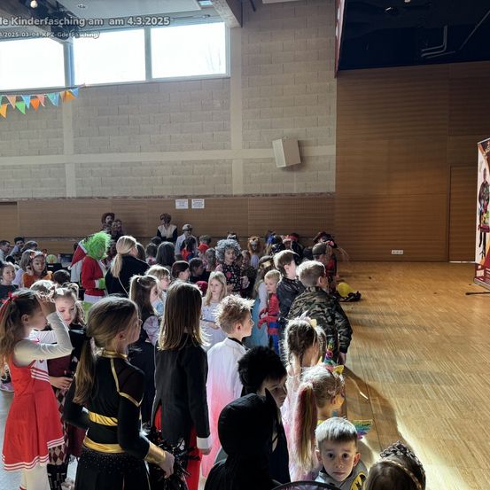Eine Menschenmenge von Kindern in Kostümen versammelt sich drinnen, wahrscheinlich für eine Feier. Der Raum hat Holzböden, eine Wand mit einem Fenster und eine Bühne. Einige Kinder tragen Masken, und Dekorationen schmücken den Bereich.