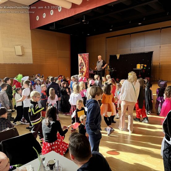 Ein Saal voller Kinder in Kostümen, die einen Mann auf der Bühne beobachten. Ein roter Banner und ein schwarzer Hintergrund mit Objekten sind vorhanden. Einige Erwachsene sind anwesend, und einige Tische mit Gegenständen sind zu sehen.