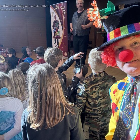 Ein Clown unterhält eine Gruppe von Kindern. Hinter ihnen führt ein Zauberer Tricks vor. Ein Plakat mit 'Magic Junior' ist sichtbar.
