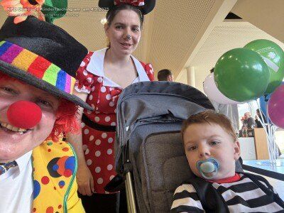 Ein Clown mit einem mehrfarbigen Hut und Gesichtsbemalung steht neben einer Frau in einem roten Punktekleid. Ein Kind mit einem Schnuller sitzt in einem Kinderwagen, und bunte Luftballons sind im Hintergrund.